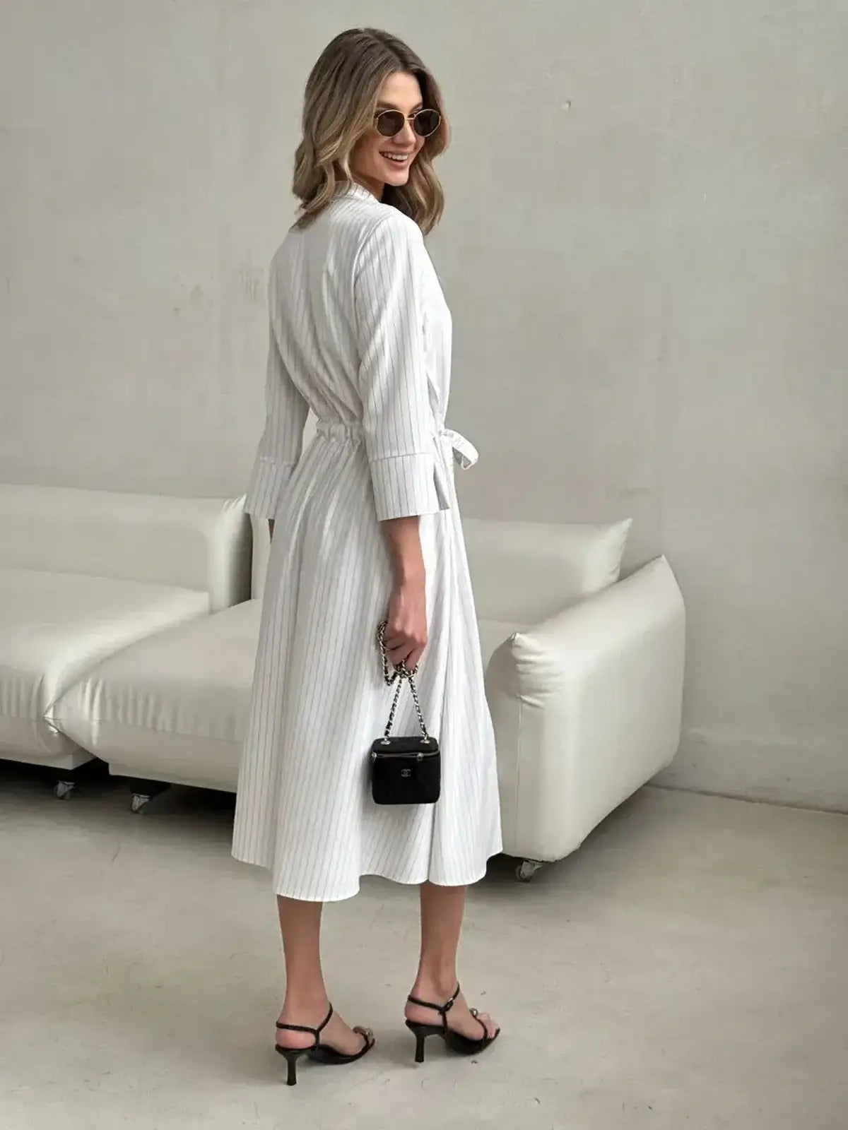 White pinstripe midi dress with waist tie, elegant for weddings or casual outings, styled with sunglasses and a small handbag.