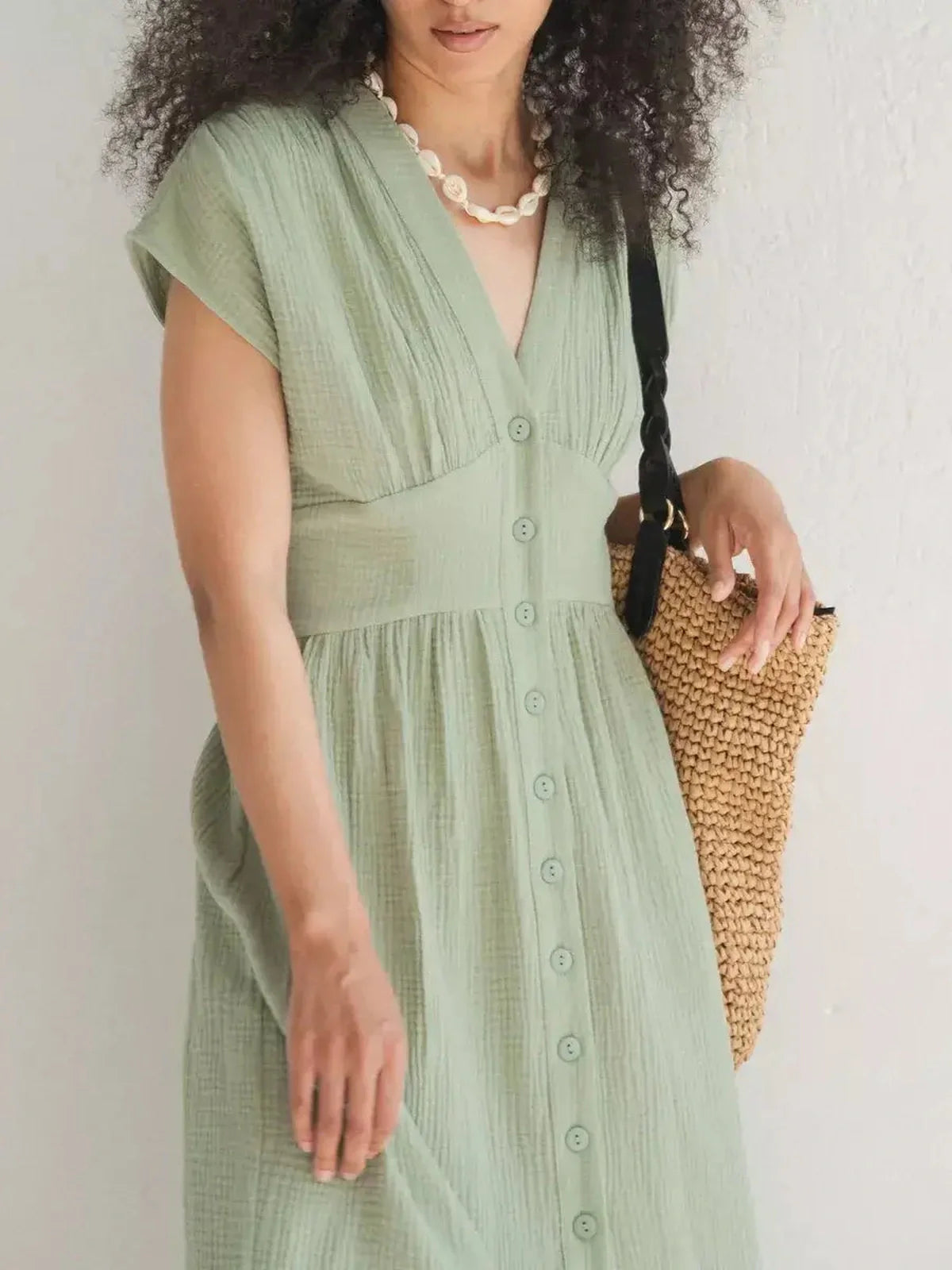Woman wearing a khaki Verdana midi dress with corset-inspired fit, accessorized with a woven bag and pearl necklace.