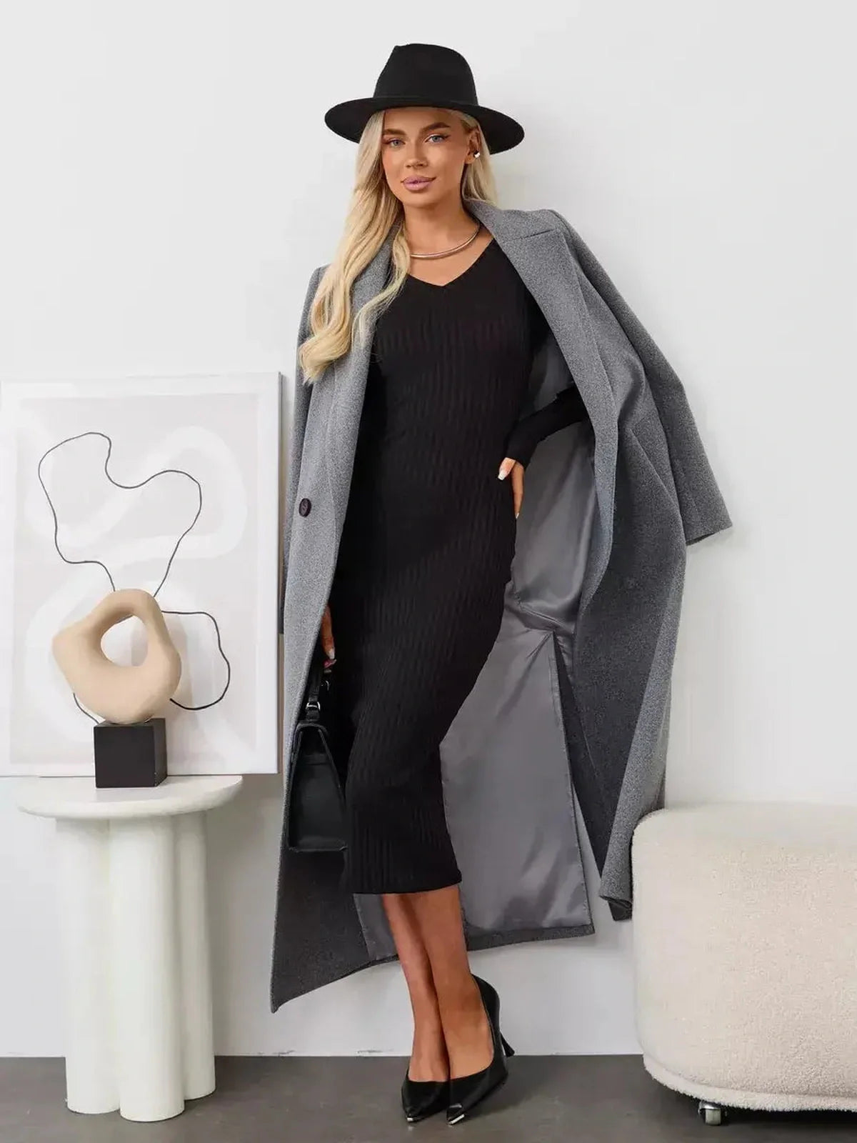Woman in a black Muse Midi Dress and gray coat, showcasing modern elegance with a stylish hat and chic accessories.
