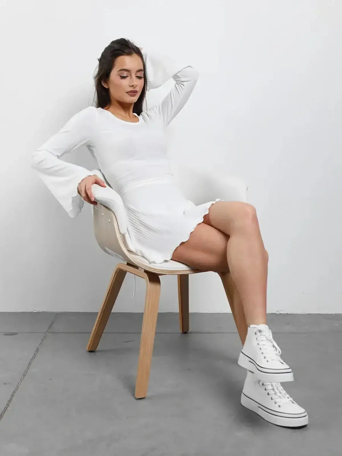 Model wearing a Luxe Lace-Up white mini dress with flared sleeves and sneakers, sitting on a chair.