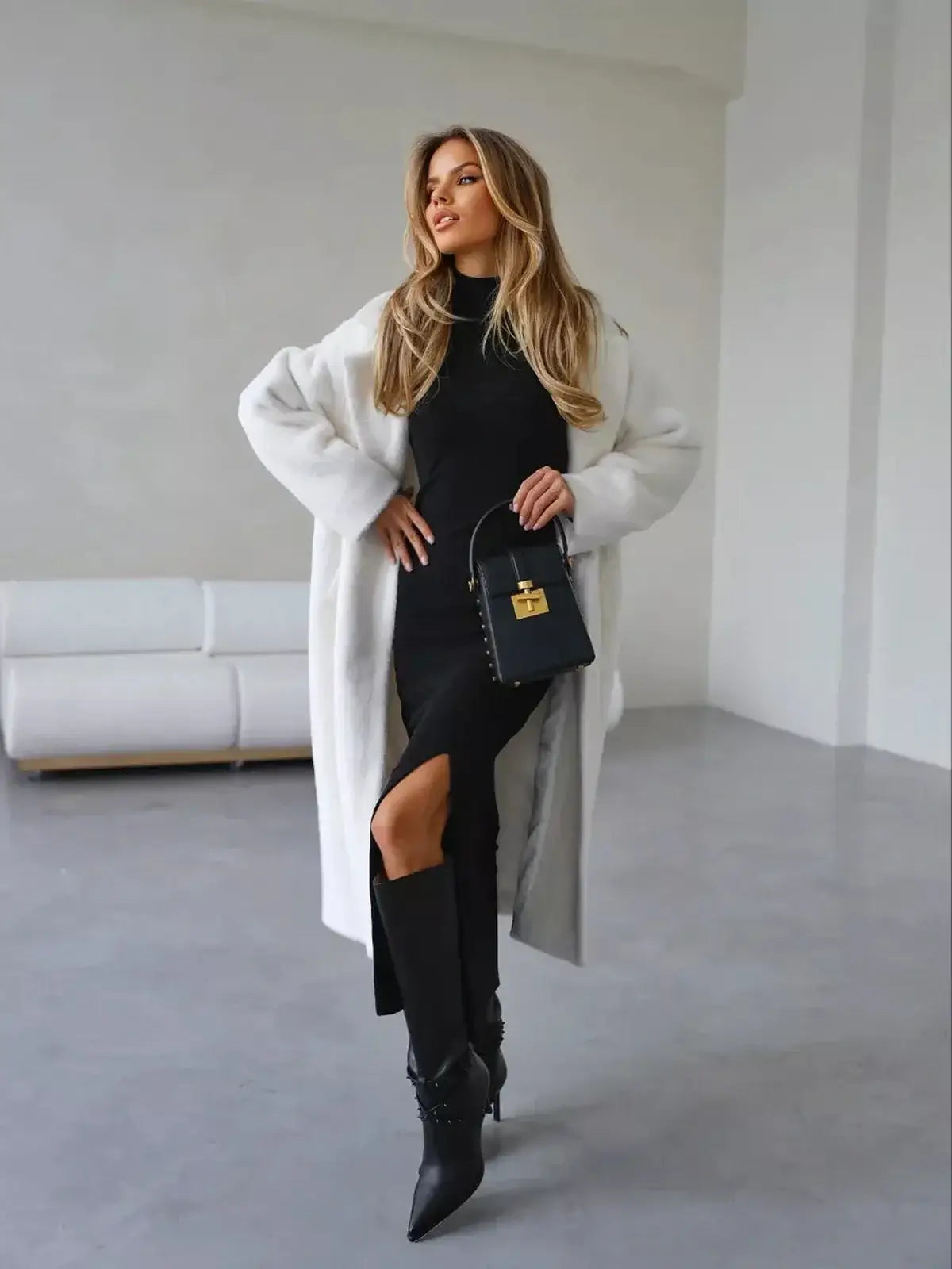 Elegant woman in a black midi dress with a high slit, styled with a white coat and chic handbag, perfect for sophisticated occasions.