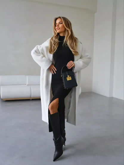 Elegant woman in a black midi dress with a high slit, styled with a white coat and chic handbag, perfect for sophisticated occasions.