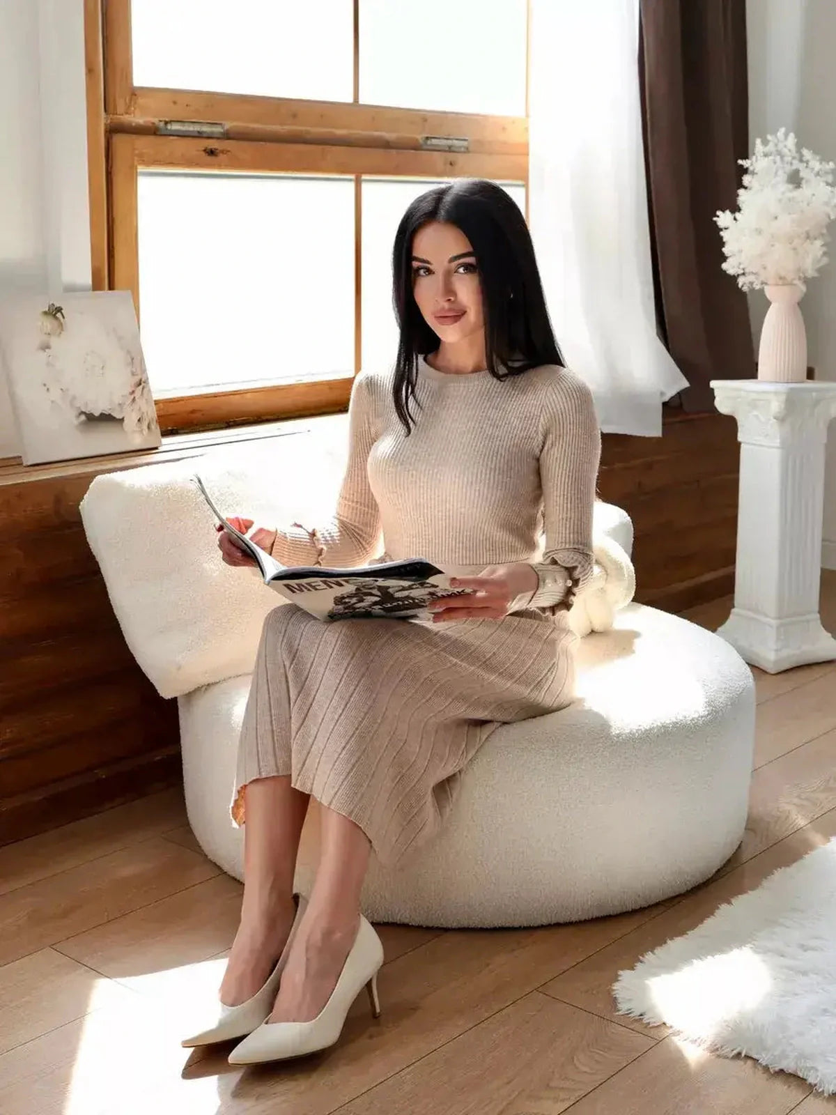 Woman in a ribbed knit midi dress sits elegantly, holding a magazine in a bright, cozy indoor setting.