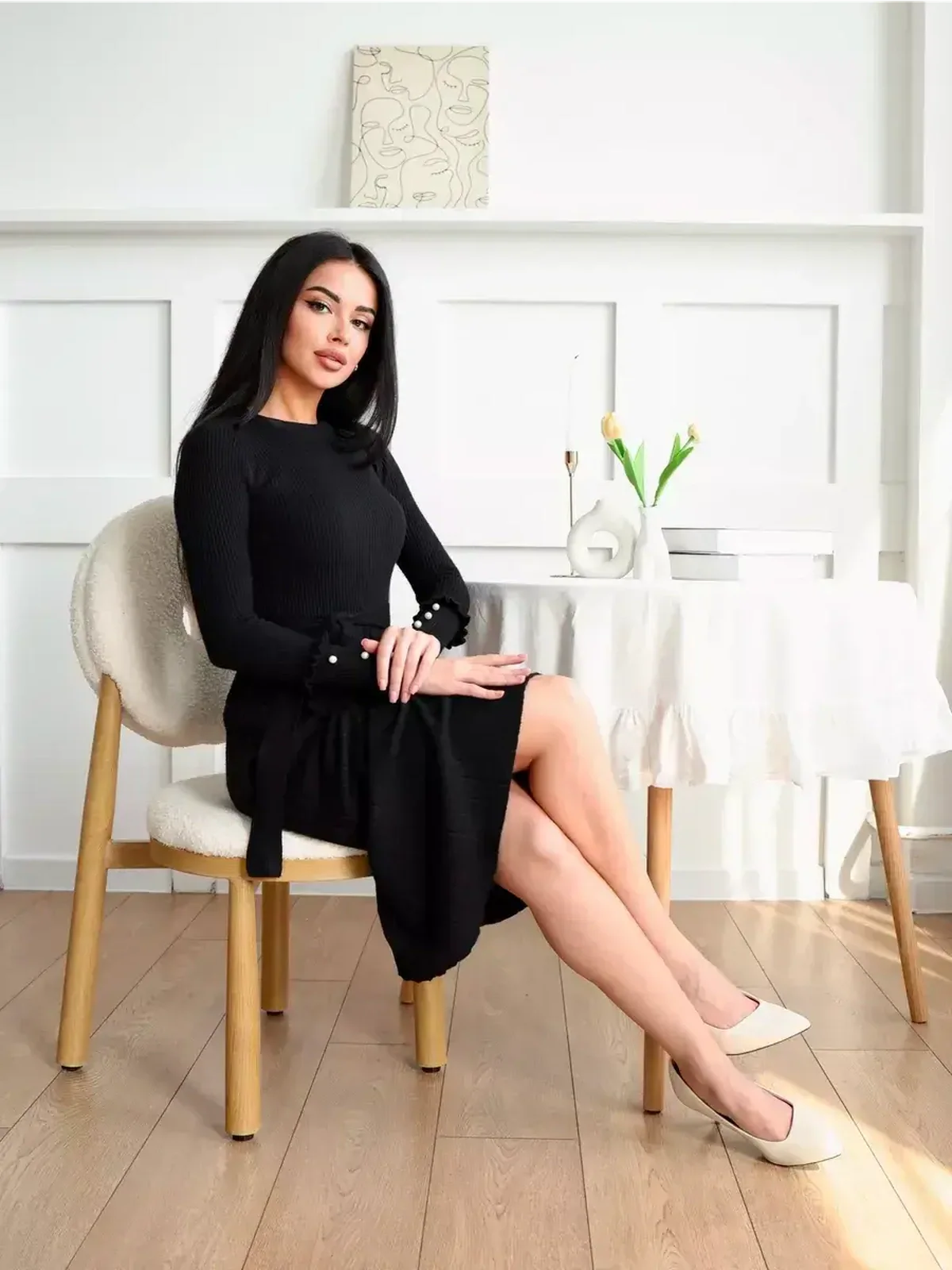 Elegant model in a black ribbed midi dress with pearl details, seated in a stylish interior.