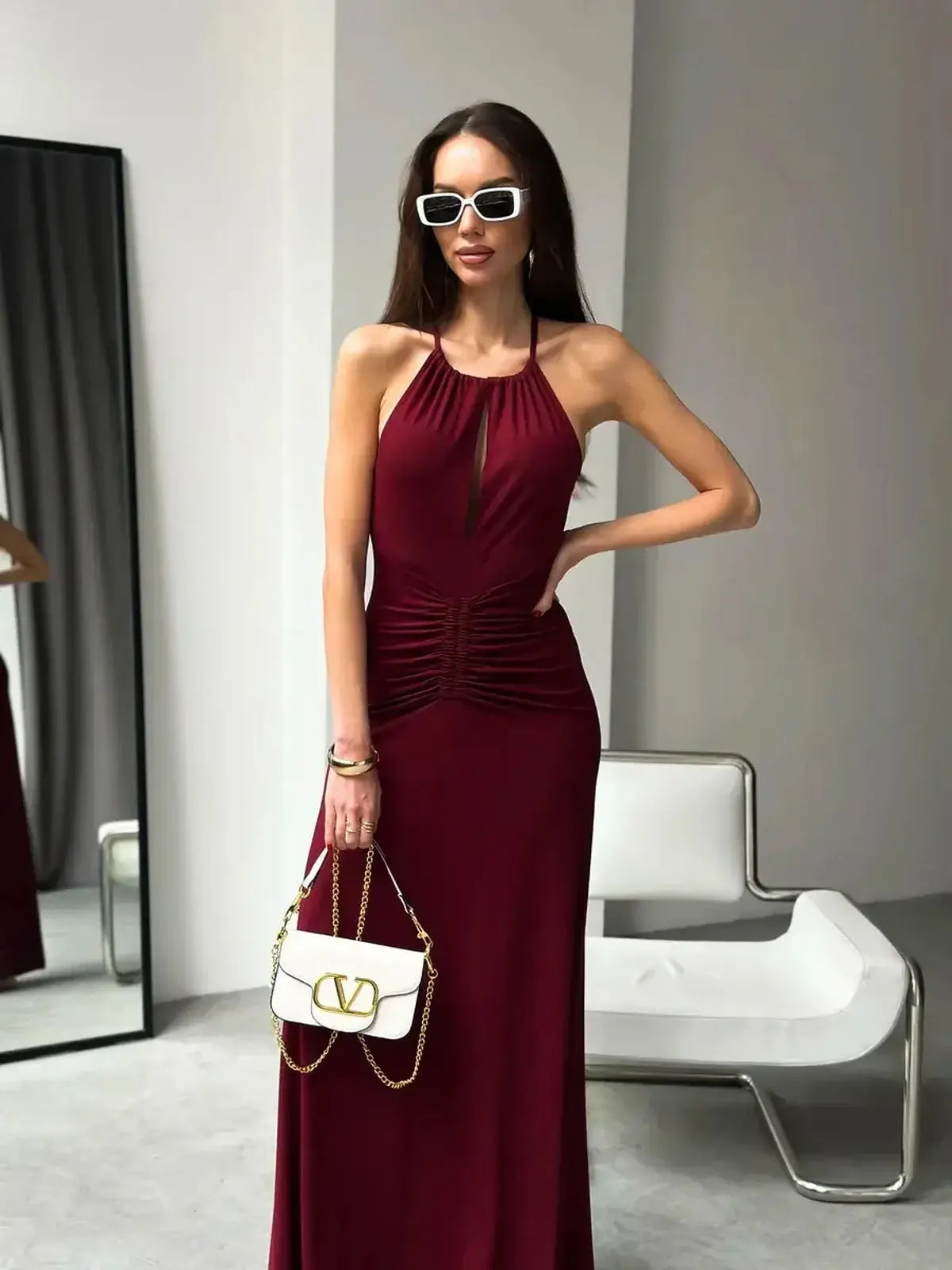 Model wearing a burgundy Maxi Dress Élan with draped halter design and ruched waist, paired with stylish sunglasses and a handbag.