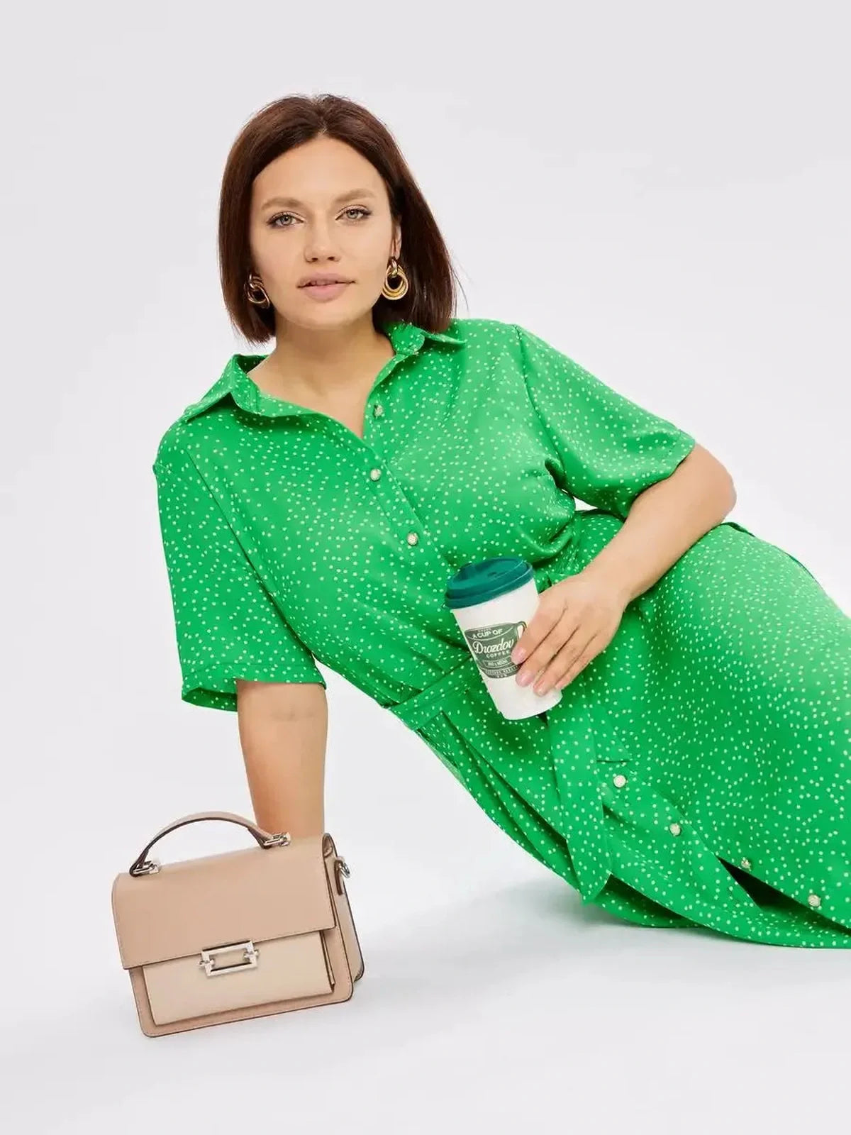 Model wearing a vibrant green Garden Dot Midi Dress with white dots, holding a coffee cup and sitting with a beige handbag.