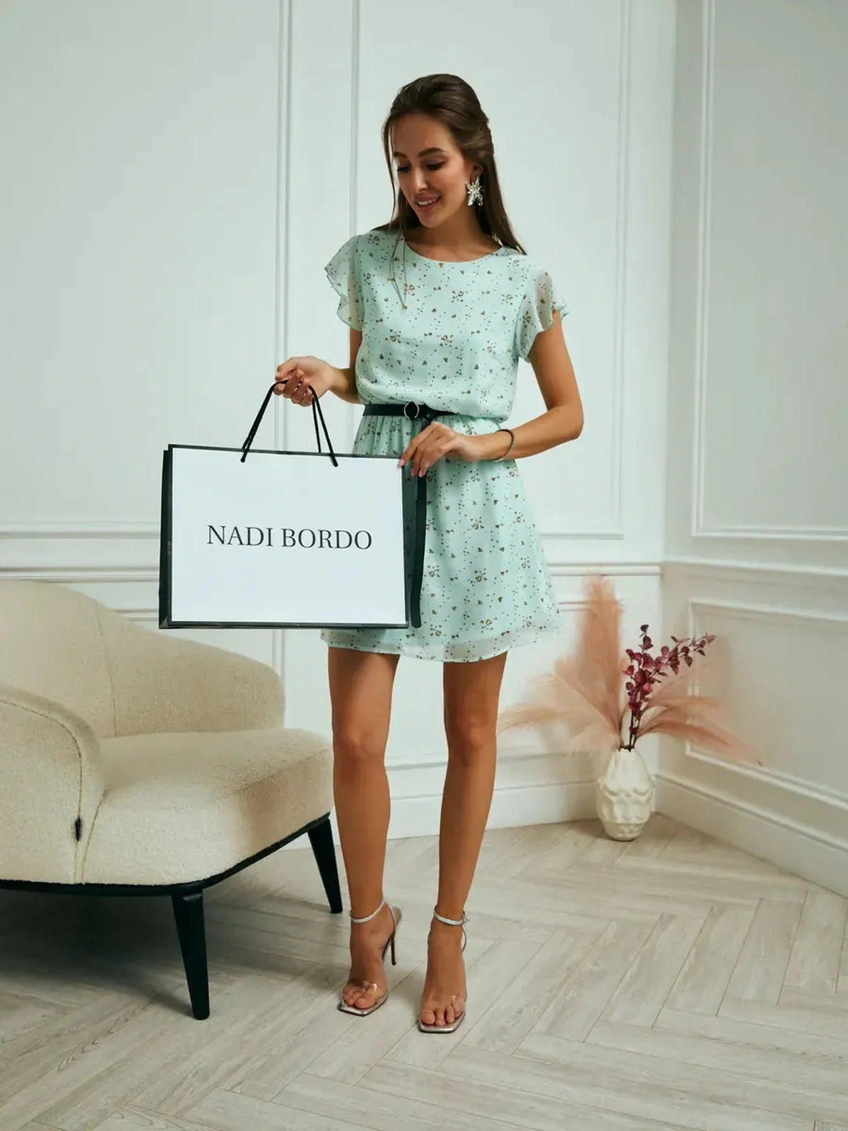 Model showcasing Mint Glow Mini Dress with fluttery sleeves and cinched waist, holding shopping bag in a stylish setting.