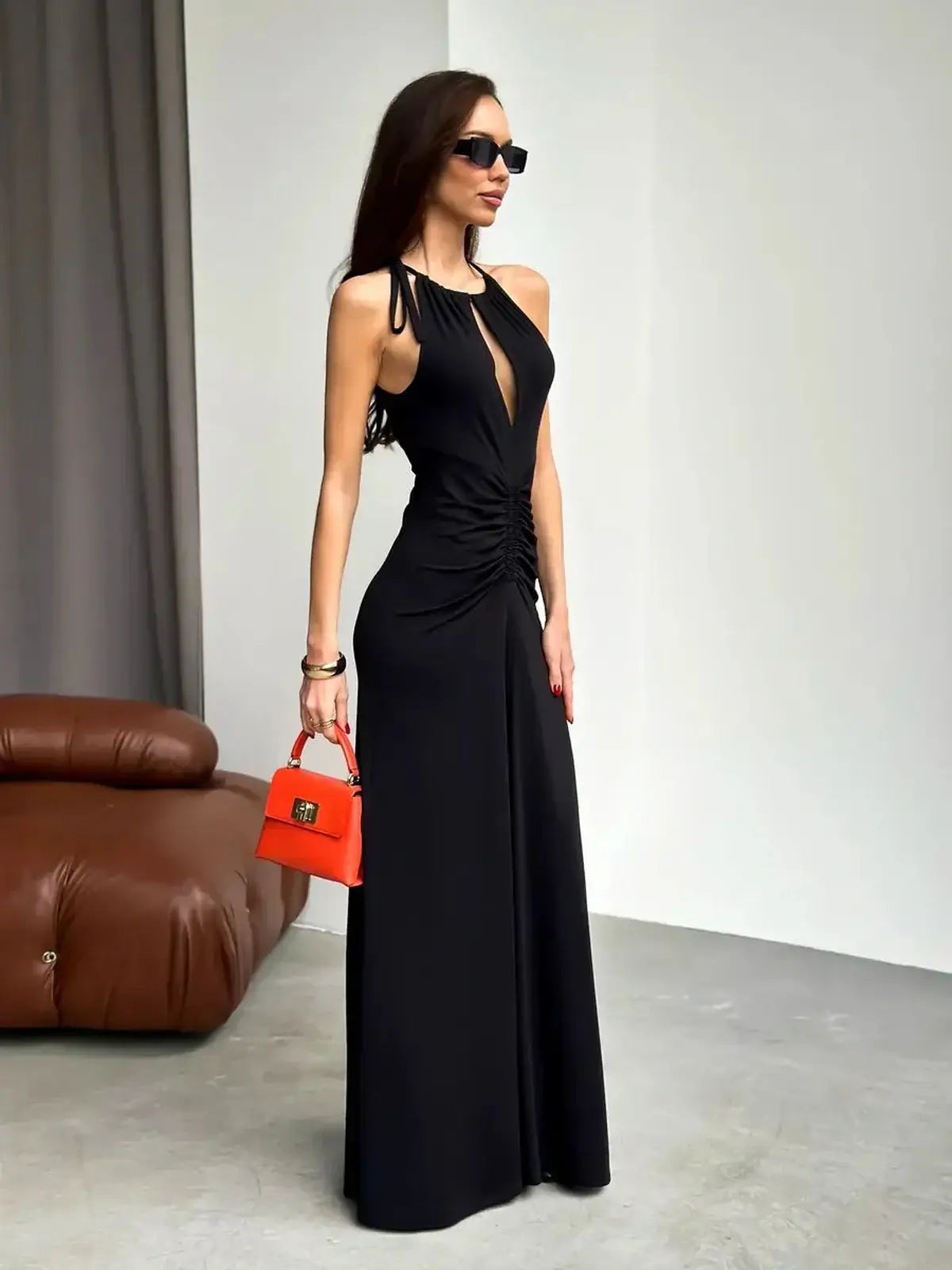 Elegant black maxi dress with ruched halter design, featuring a front slit and paired with a stylish orange handbag.