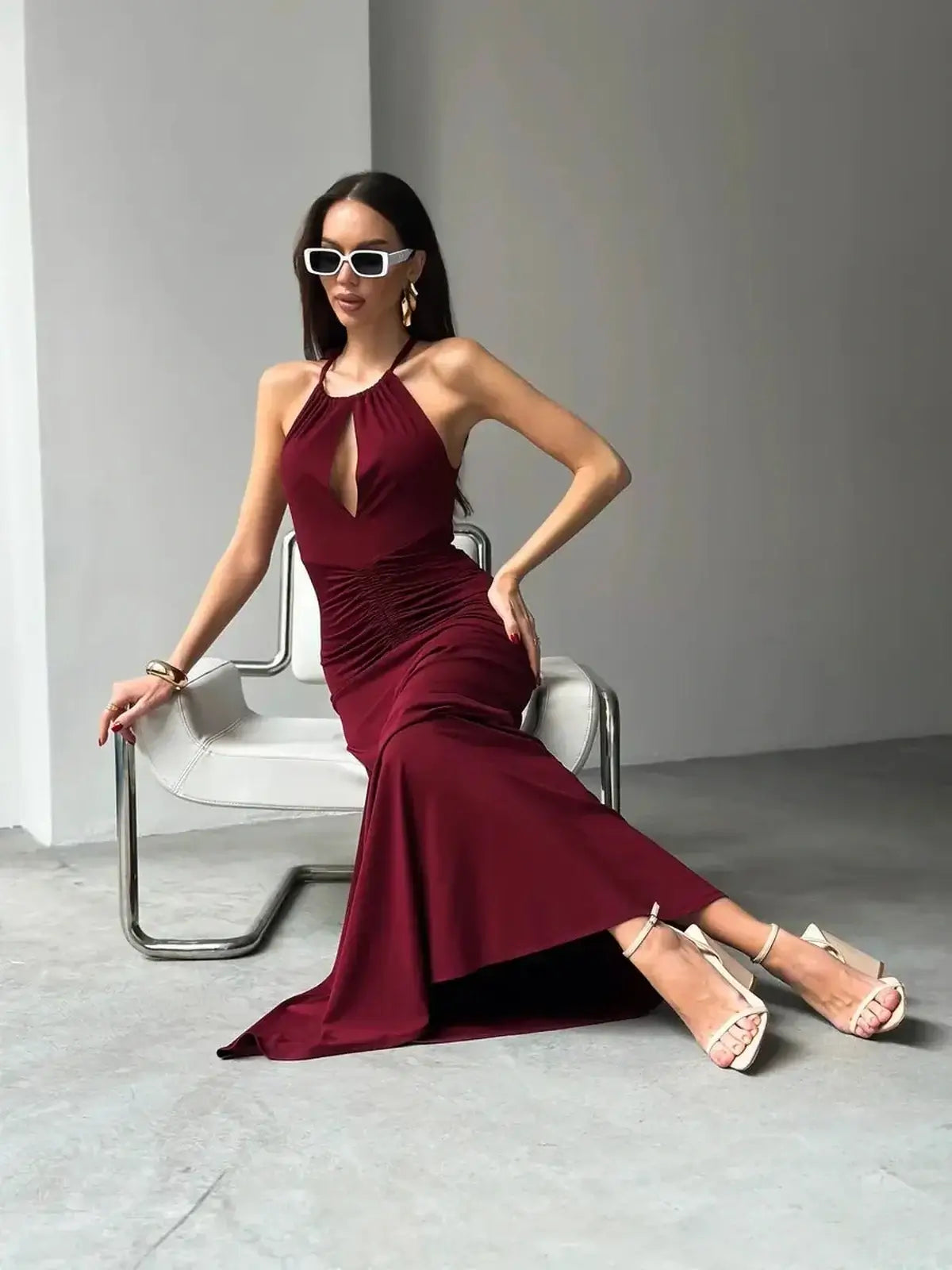 Model in a burgundy halter maxi dress with a keyhole front, sitting stylishly in a modern space, perfect for formal events.