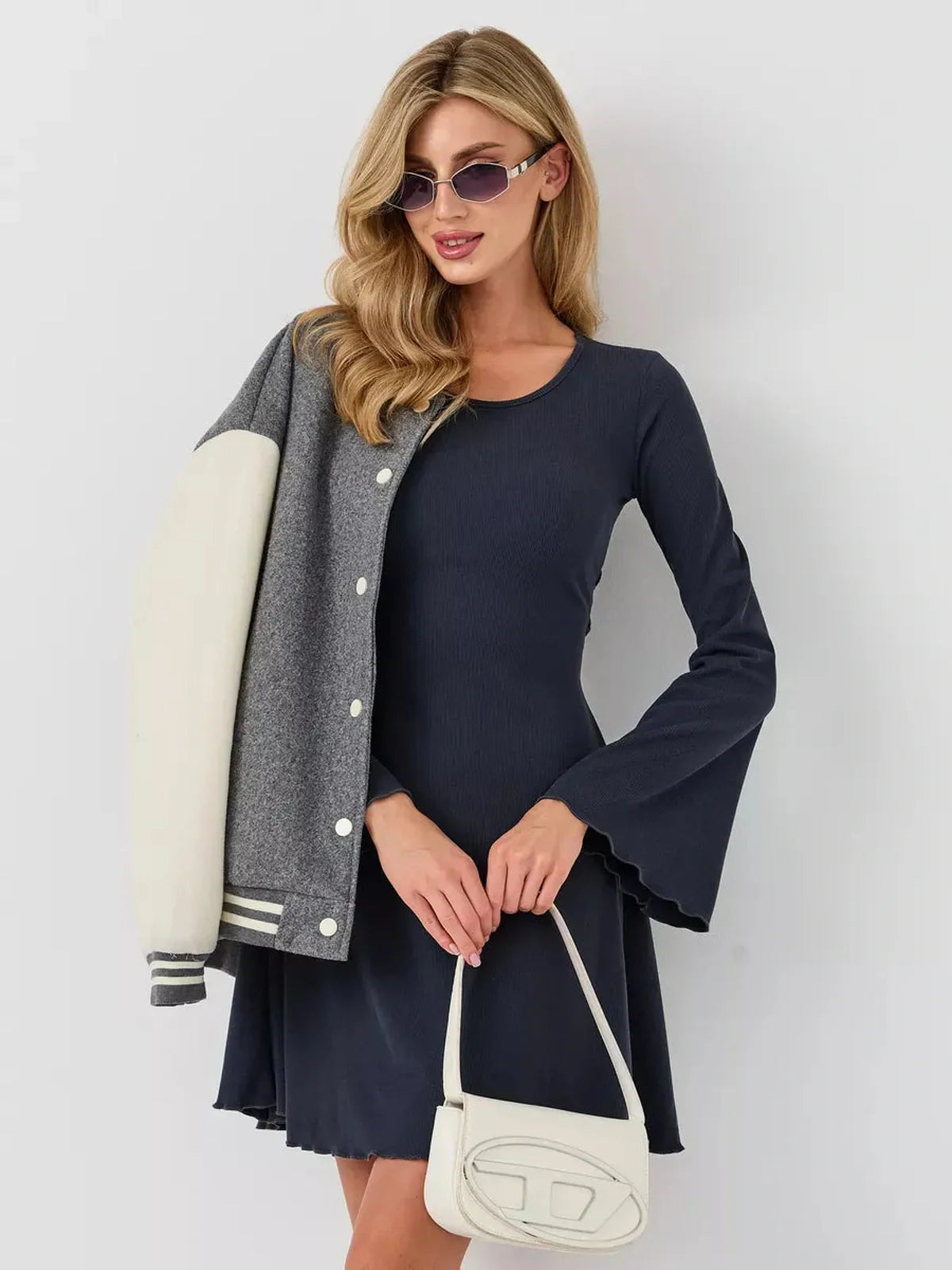 Model wearing a navy mini dress with bell sleeves, paired with a grey varsity jacket and stylish sunglasses.