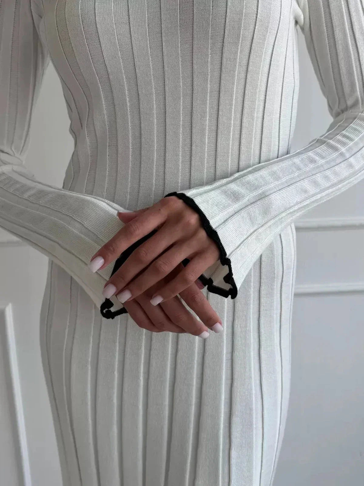 Close-up of a woman's hands in a ribbed ivory maxi dress, showcasing elegant long sleeves with delicate black ruffle trim.