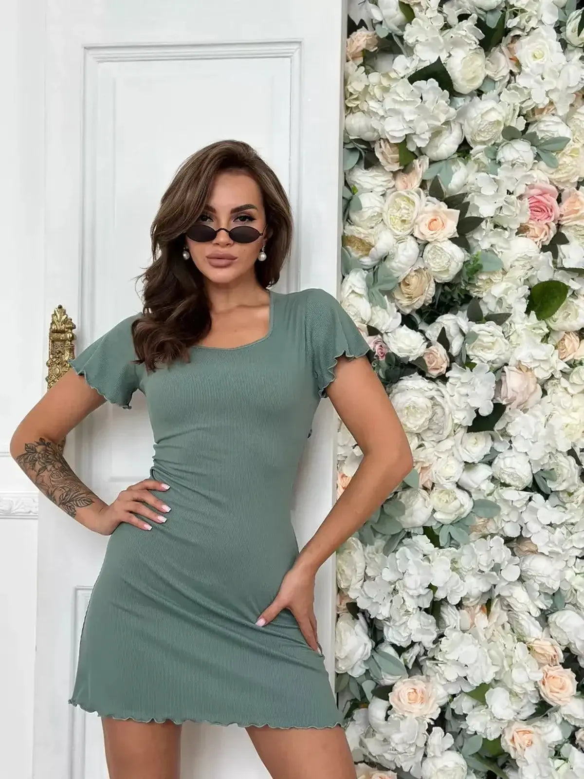 Model showcasing the Sage Aura Mini Dress with flutter sleeves against a floral backdrop, perfect for summer or wedding guest attire.