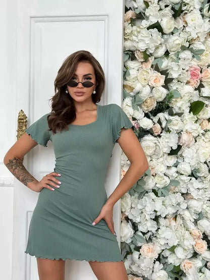 Model showcasing the Sage Aura Mini Dress with flutter sleeves against a floral backdrop, perfect for summer or wedding guest attire.