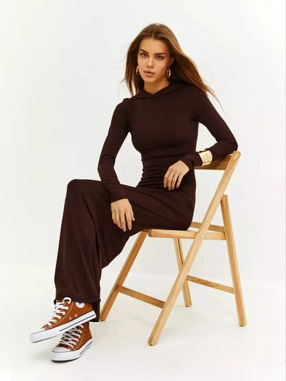 Woman in a brown Sculpt Maxi Dress with hood, styled casually with sneakers, seated on a wooden chair.