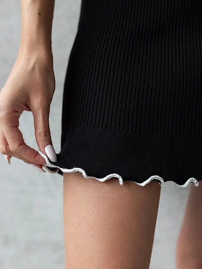 Close-up of a woman's hand adjusting the flouncy hem of a black ribbed mini dress with white contrast edging.