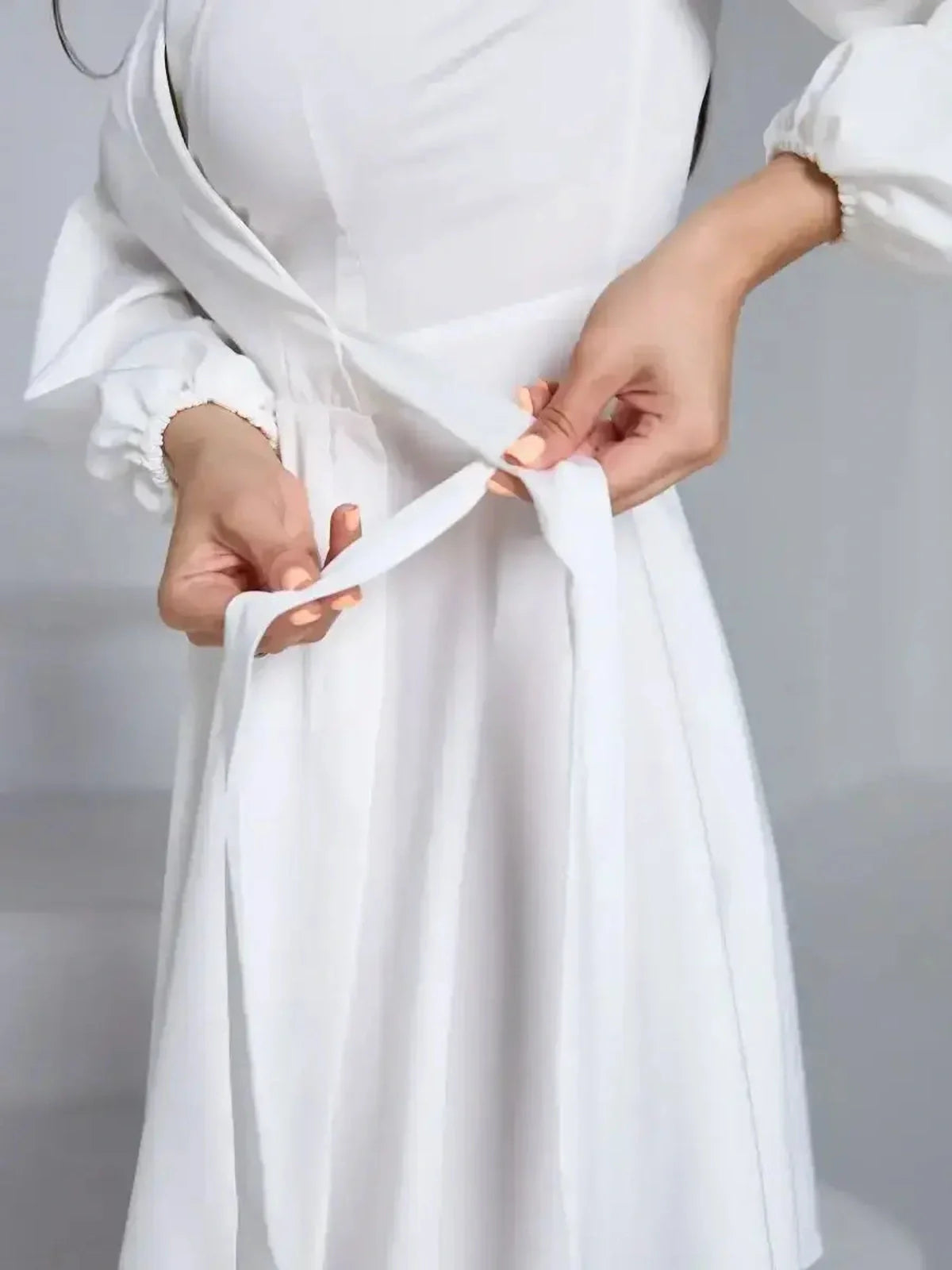 Close-up of a woman tying the adjustable waist tie on a white dress, showcasing its elegant wrap-front design.