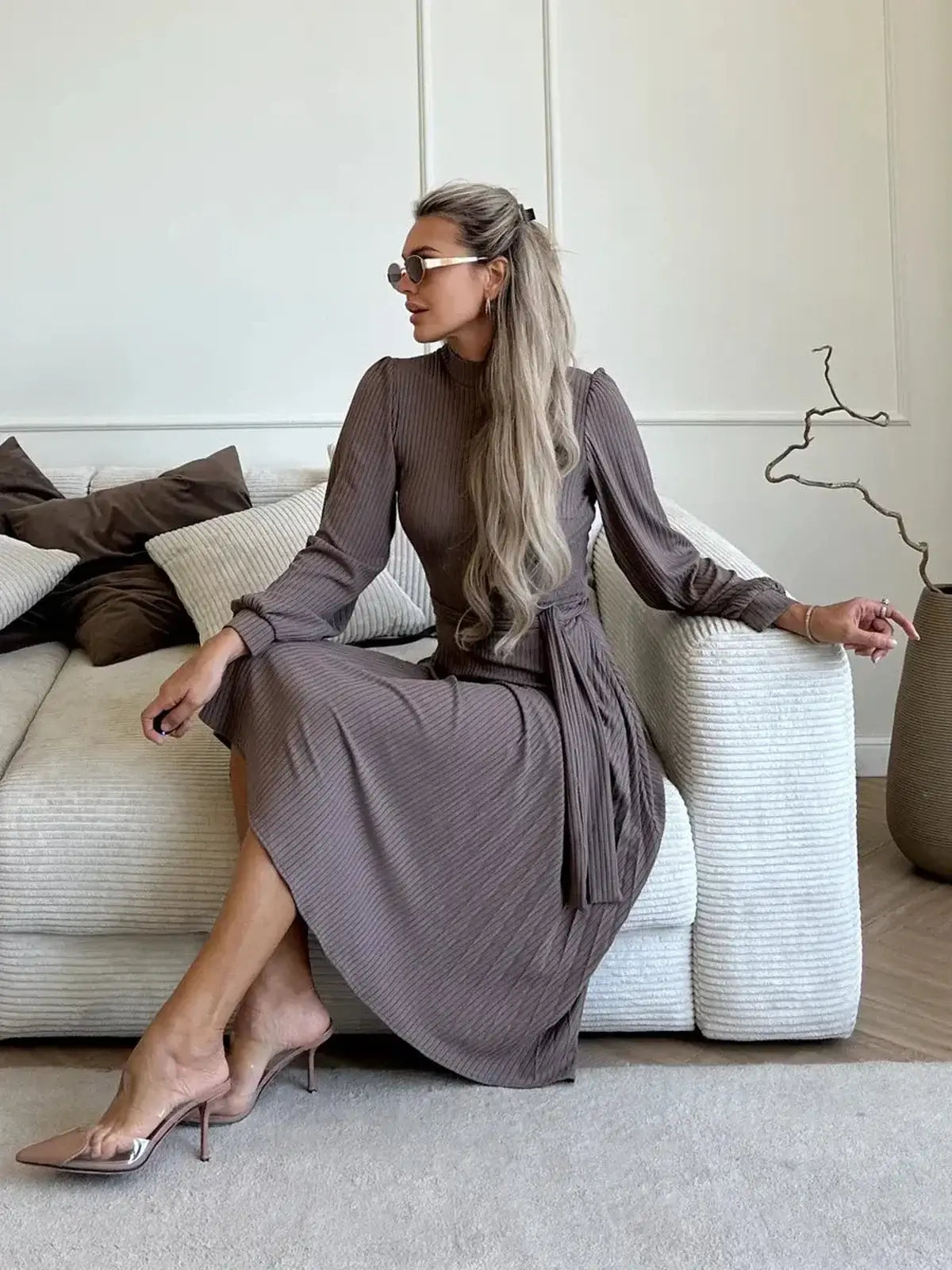 Woman in a ribbed Plissé Charm Midi Dress sits on a sofa, showcasing its elegant fit and pleated skirt in a rich coffee hue.