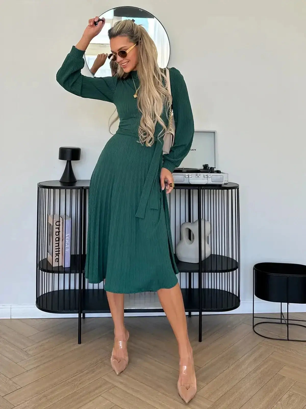 Woman wearing an elegant emerald midi dress with pleats, styled with sunglasses in a chic indoor setting.