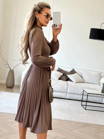 Model wearing a Plissé Charm Midi Dress in coffee color, showcasing a pleated skirt and elegant fit in a stylish indoor setting.