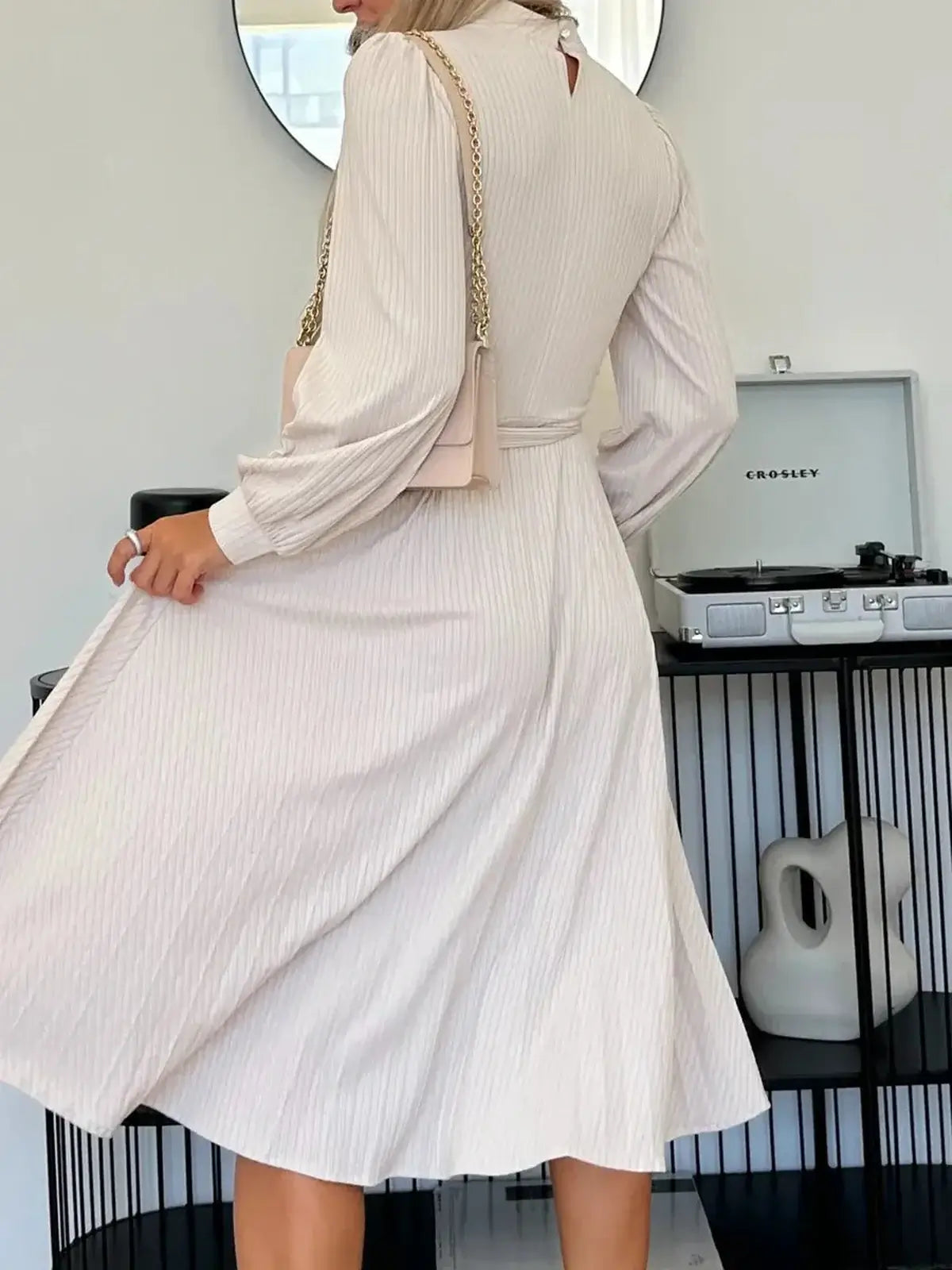 Back view of a woman in an ivory midi dress with ribbed texture, featuring flowing pleats and subtle puffed sleeves.