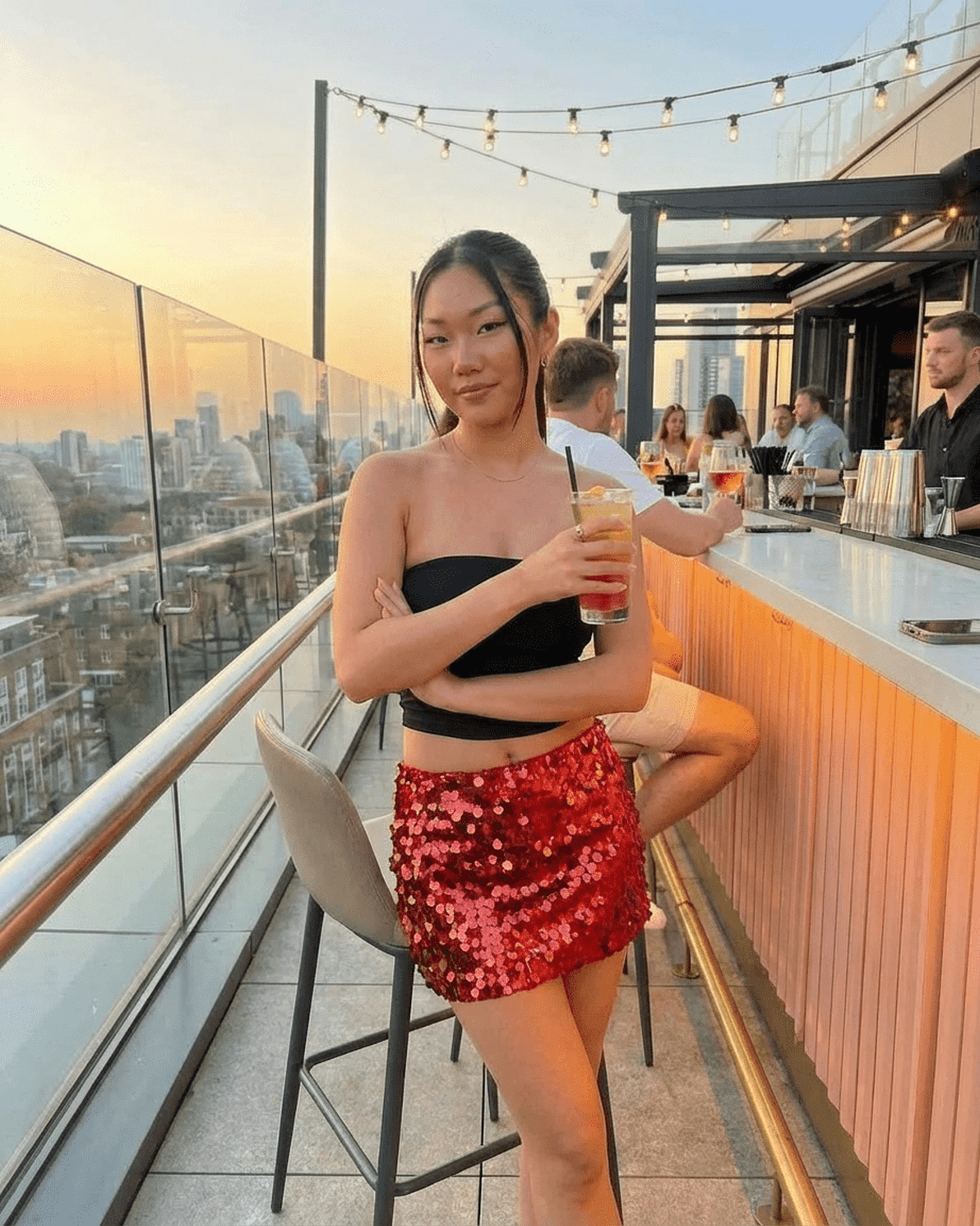 Model wearing Slimona Flamenco Mini Skirt with red sequins, enjoying a cocktail at a rooftop bar during sunset.