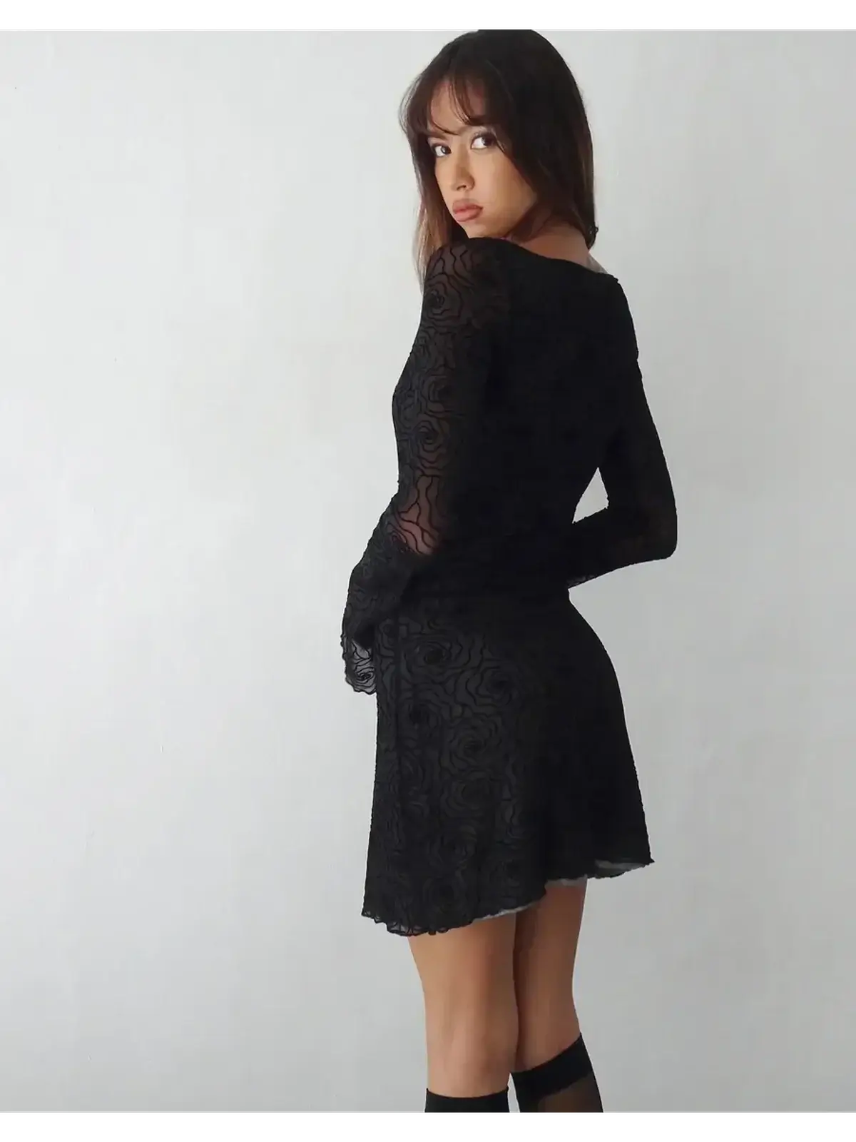 Model wearing a fitted black mini dress with long sleeves, showcasing a floral lace design.