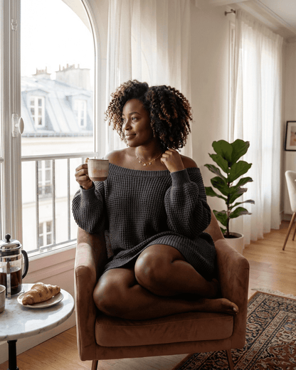 Woman in a cozy off-the-shoulder sweater enjoying coffee at home, showcasing the Ember Mini Dress from Slimona.
