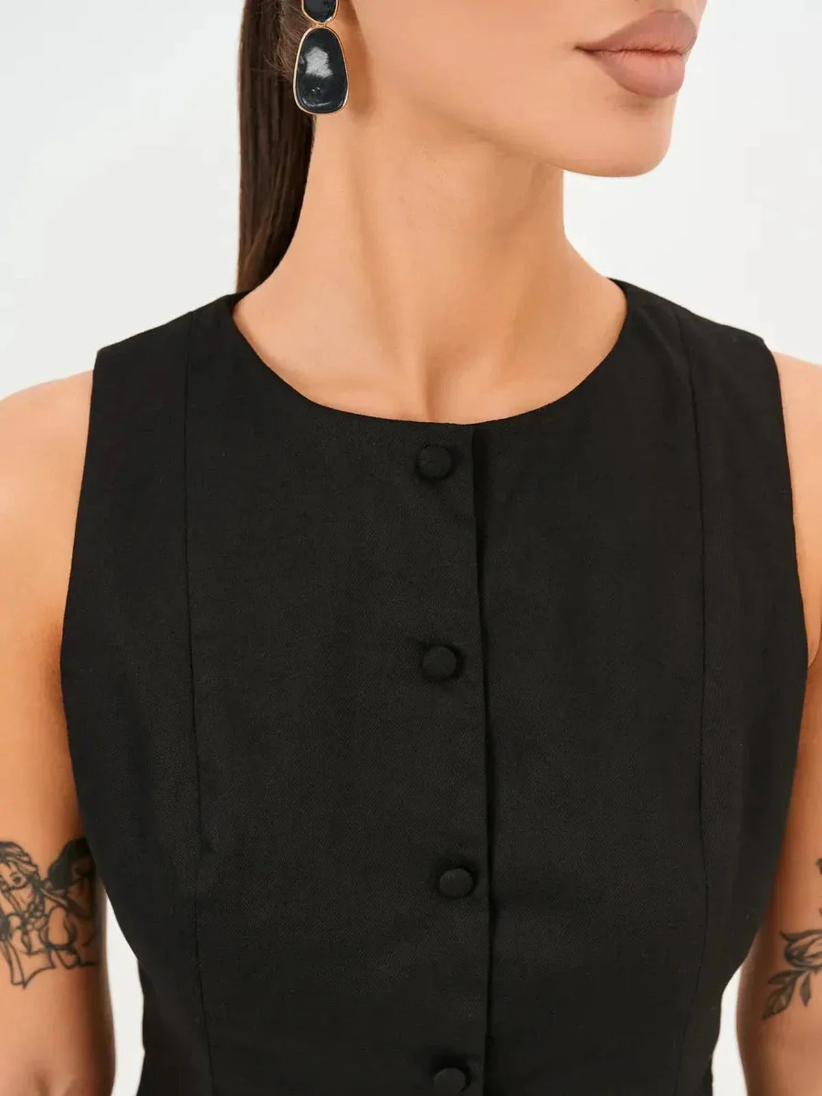 Close-up of a model wearing a black sleeveless dress with a round neckline and button detail, showcasing modern elegance.