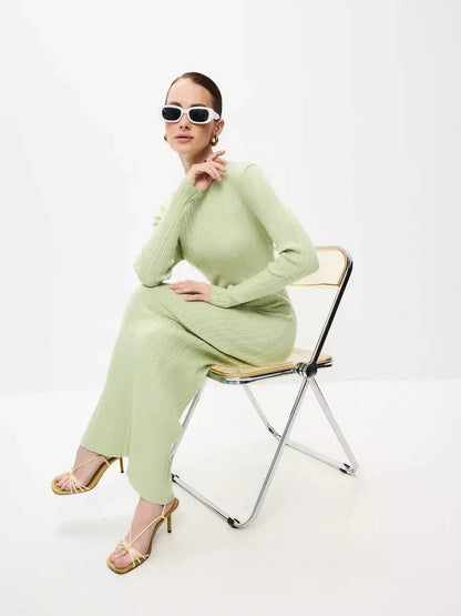 A model wearing the Celery Luxe Maxi Dress in light green, posing elegantly on a chair with stylish sunglasses.