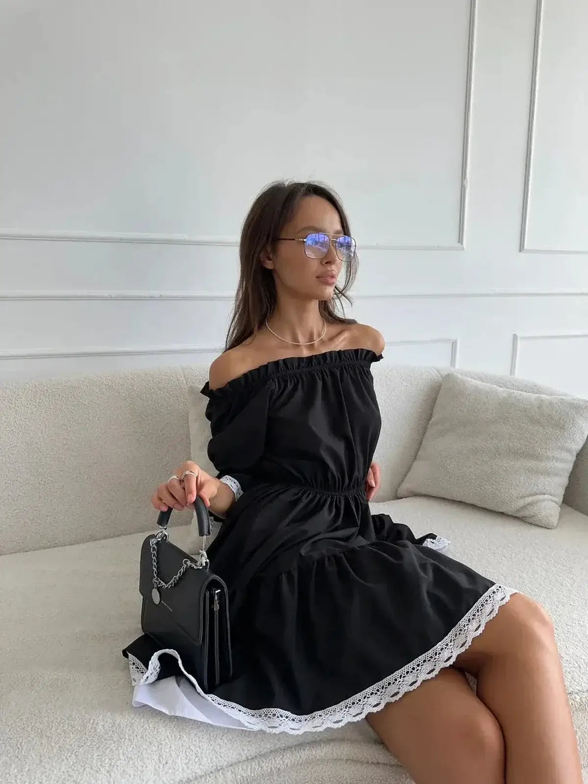 Woman in a black off-shoulder mini dress with lace trim, seated stylishly on a couch.