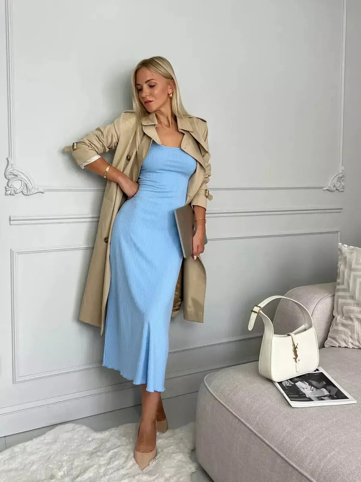 Stylish woman in a sky blue midi dress paired with a beige trench coat, posing elegantly by a modern sofa.