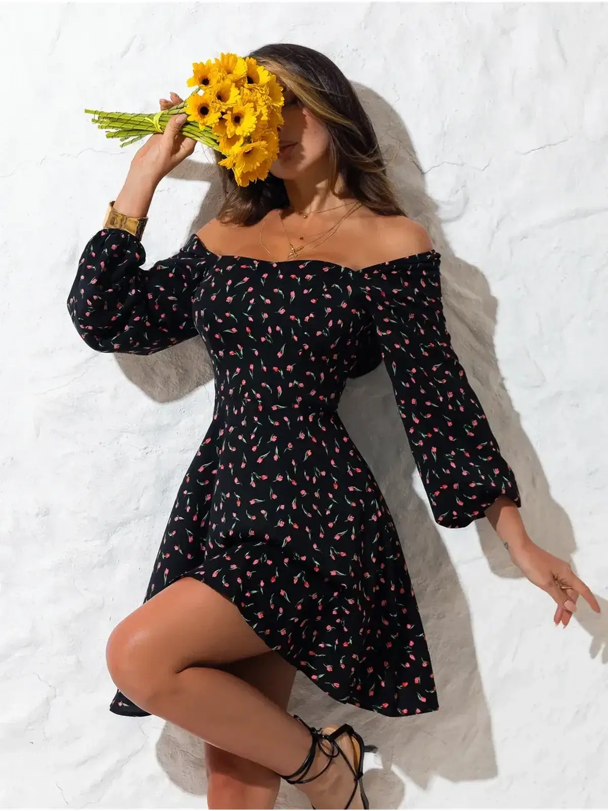 A woman in a black floral mini dress with puffed sleeves, holding sunflowers against a white wall, ideal for summer occasions.