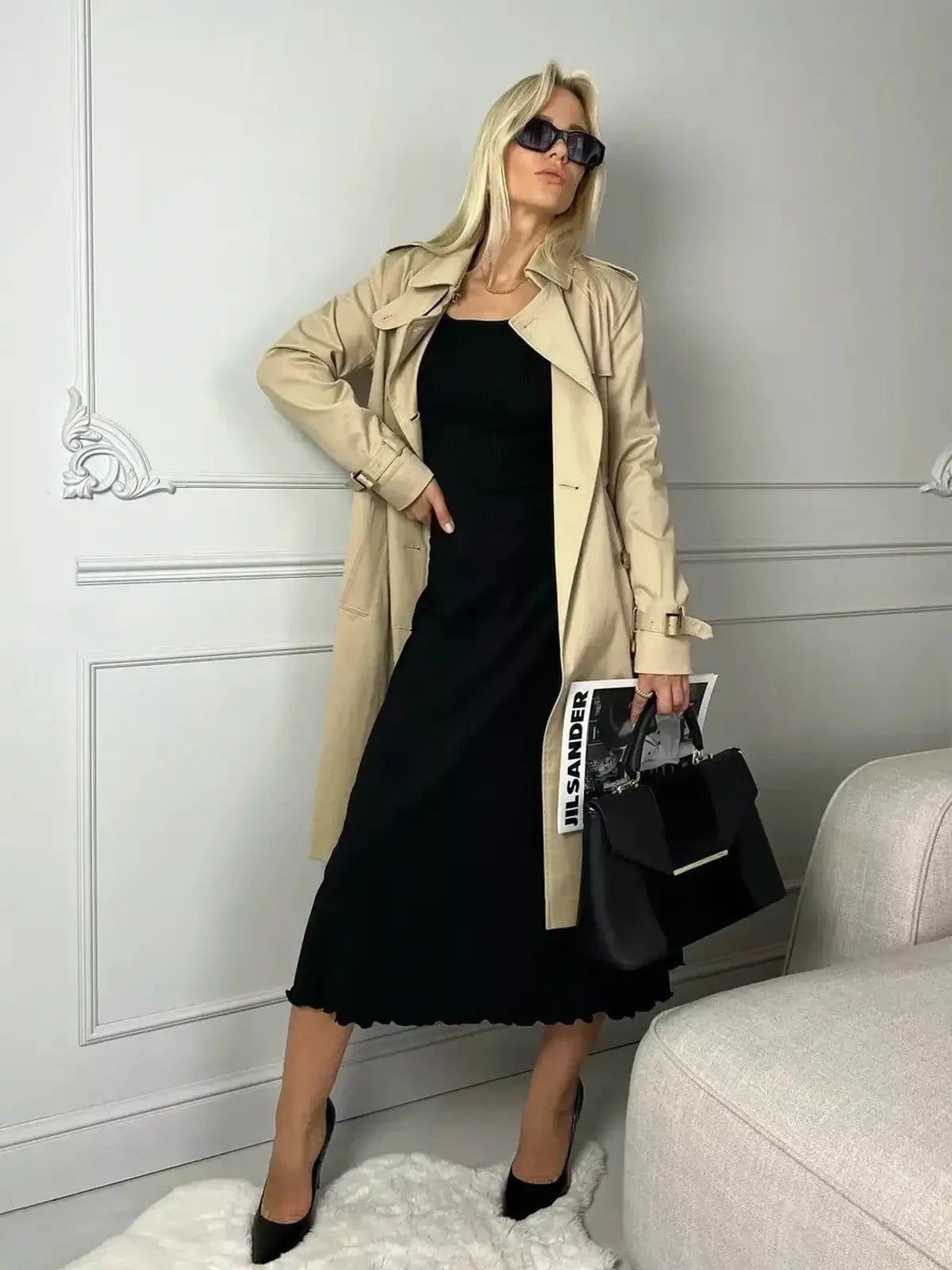 Stylish woman in a black midi dress and beige trench coat, accessorized with sunglasses and a handbag, poised for a chic look.