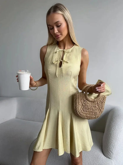 Model showcasing a Lemon Flare sleeveless mini dress in yellow, perfect for summer, styled with a woven bag and drink.