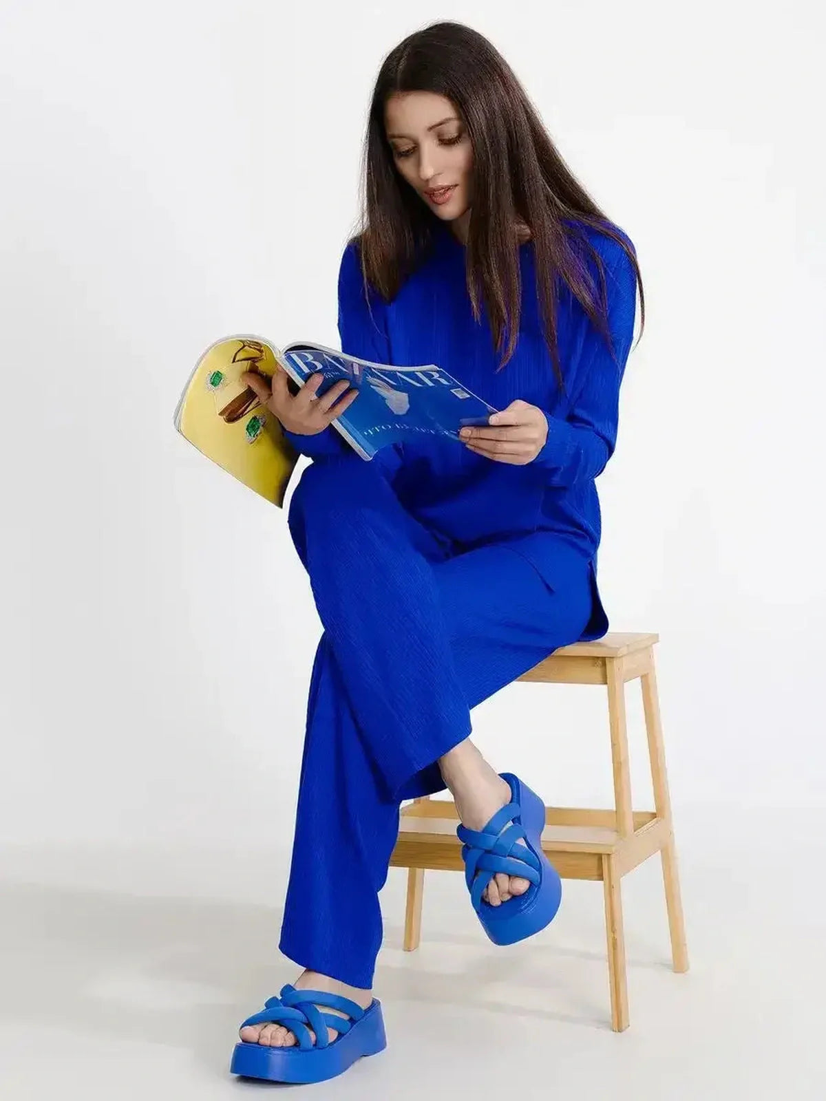 Woman in electric blue lounge set reading a magazine while sitting on a stool, showcasing relaxed elegance and style.