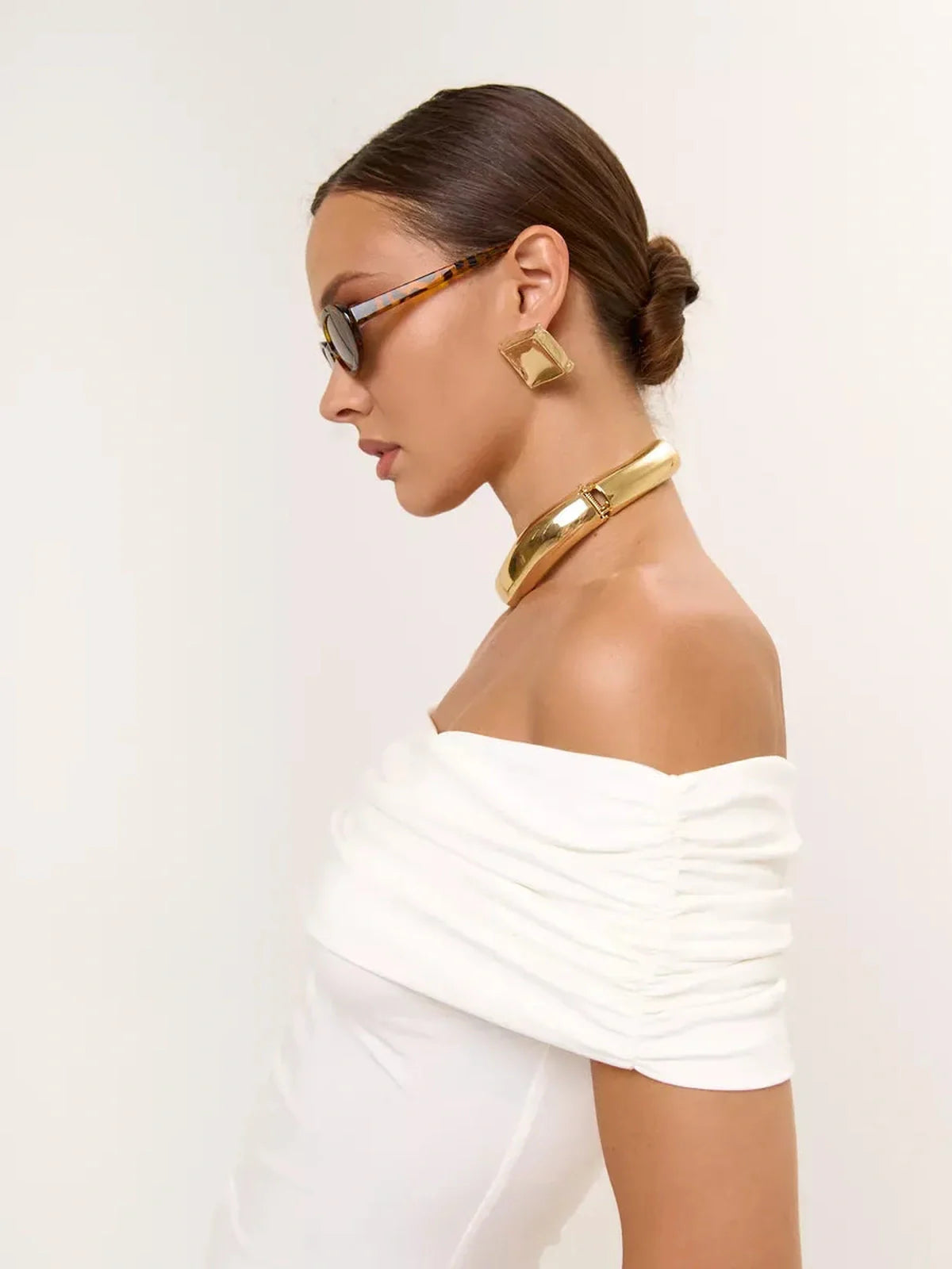 Model wearing off-shoulder ivory mini dress with gold accessories and sunglasses, showcasing elegant summer fashion.