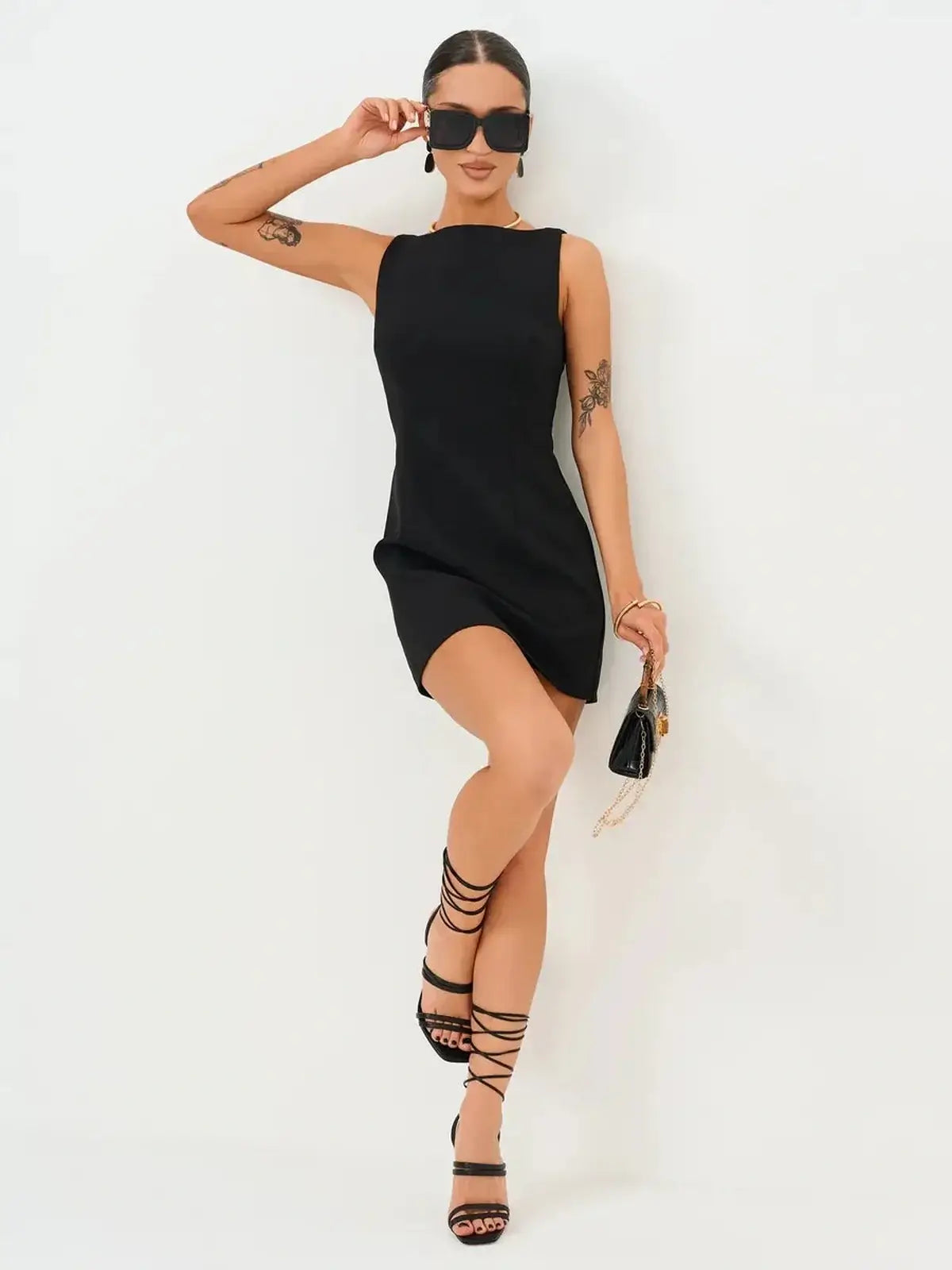 Model wearing a sleek black Jet Luxe Mini Dress with strappy heels and sunglasses, showcasing a trendy look.