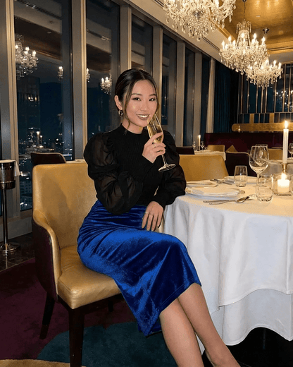 Woman wearing Azure Midi Skirt by Slimona, enjoying a drink at an elegant restaurant setting.