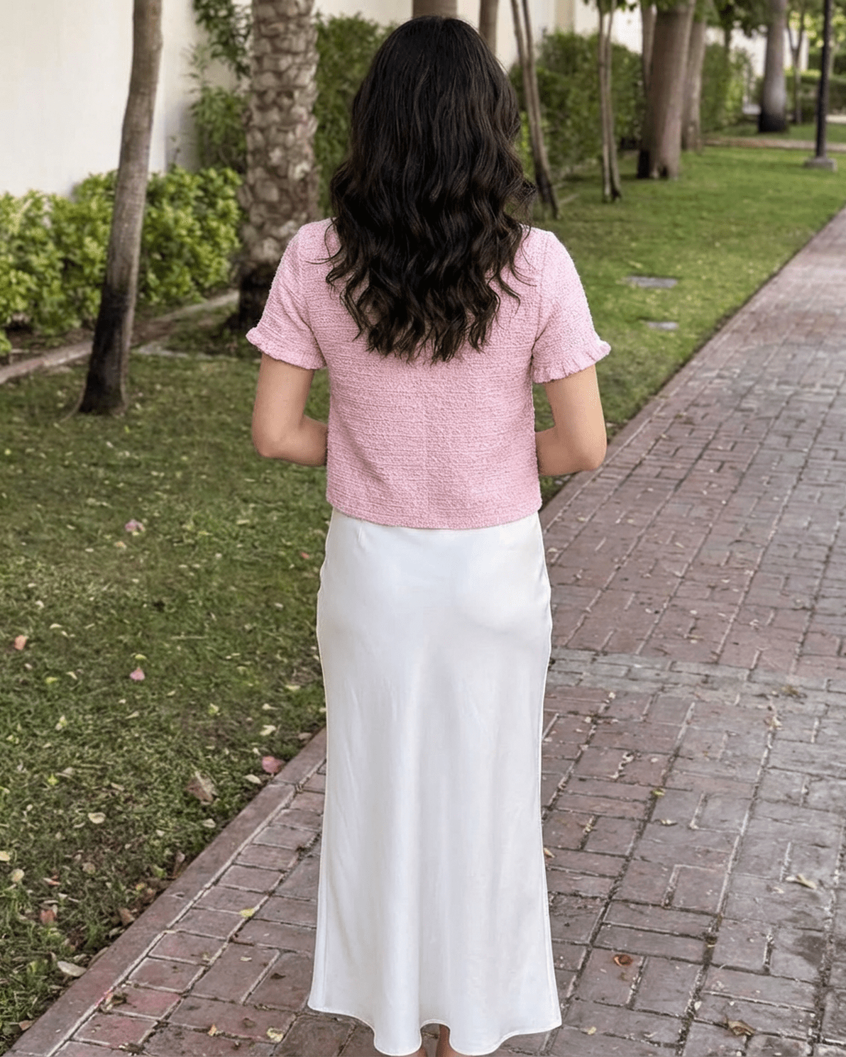 Back view of a model wearing the Slimona Camellia Cardigan in pink paired with a white skirt on a scenic pathway.