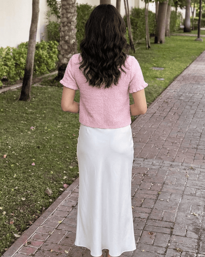 Back view of a model wearing the Slimona Camellia Cardigan in pink paired with a white skirt on a scenic pathway.