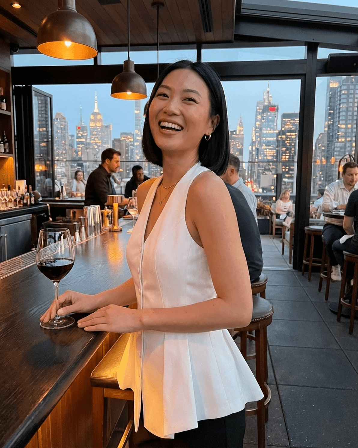Woman wearing a Celeste Blouse by Slimona, smiling at a bar with a wine glass in hand, city skyline in the background.