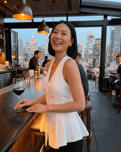 Woman wearing a Celeste Blouse by Slimona, smiling at a bar with a wine glass in hand, city skyline in the background.