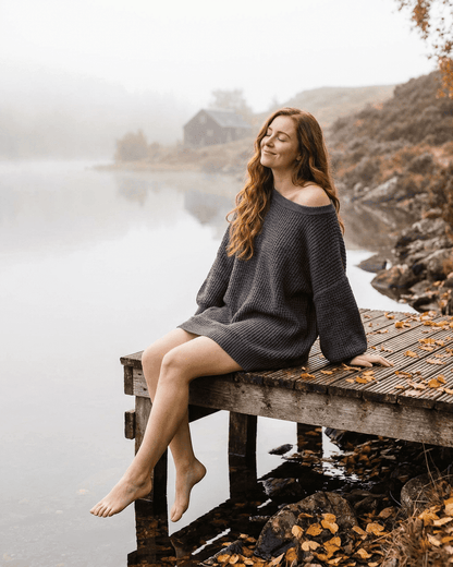 Slimona Ember Mini Dress model sitting by a lake, showcasing the cozy charcoal ribbed knit fabric and off-the-shoulder design.