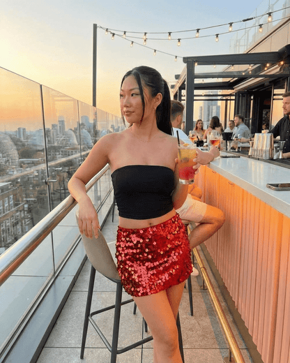 Woman wearing a Slimona Flamenco Mini Skirt, red sequins, enjoying a cocktail against a sunset backdrop.