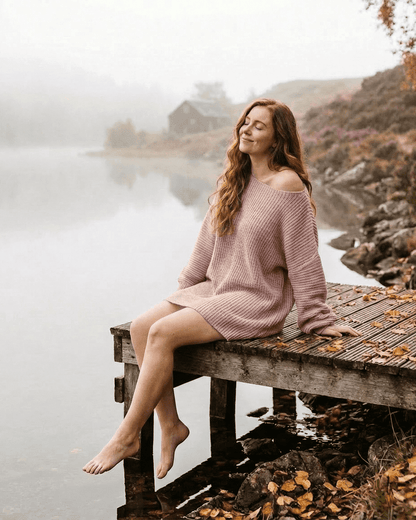 Nova Mini Dress by Slimona, showcasing a model in a soft pink ribbed knit off-the-shoulder design by a serene lake.