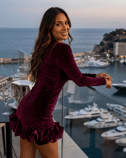 Woman wearing Velora Mini Dress by Slimona, a deep burgundy velvet bodycon dress with pleated hem, overlooking a marina.