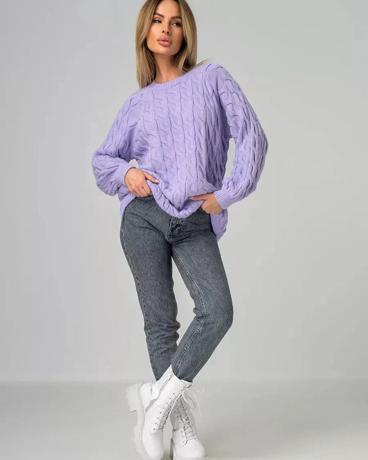 Model wearing Lyra Sweater by Slimona in oversized lavender cable knit with jeans and white boots.