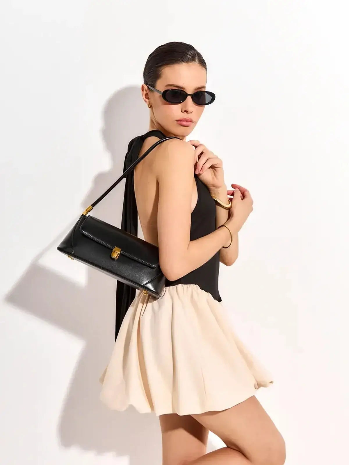 Model in a black mini dress with a beige skirt, sunglasses, and a black handbag, showcasing chic evening style.