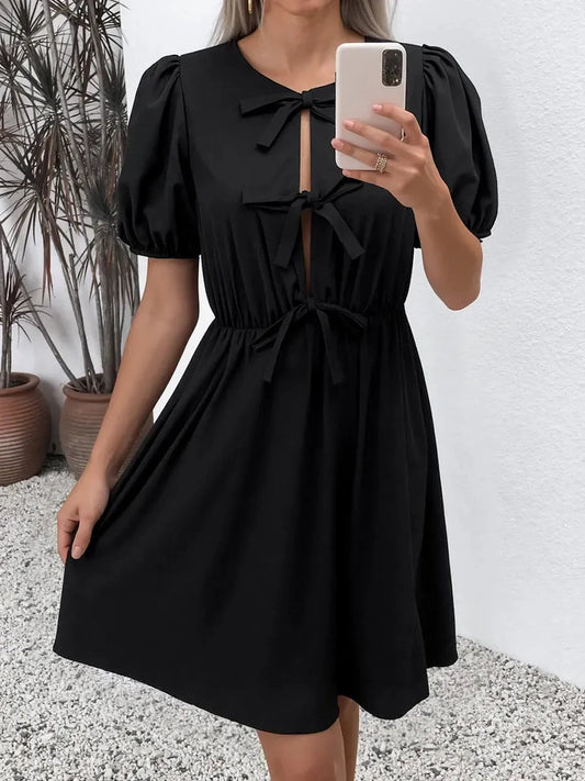 Elegant black mini dress with puff sleeves and front bow-tie closures, perfect as a wedding guest dress or for summer occasions.