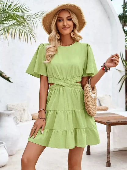 Stylish woman in a green Palm Bloom Mini Dress with ruffle skirt and flutter sleeves, accessorized with a straw hat and bag.